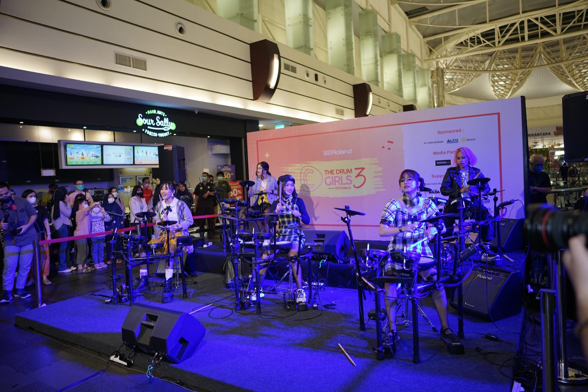 Five drummers of C The Drum Girl performing on Roland electronic kits at a mall stage, under a season three backdrop, with an audience behind the barrier.