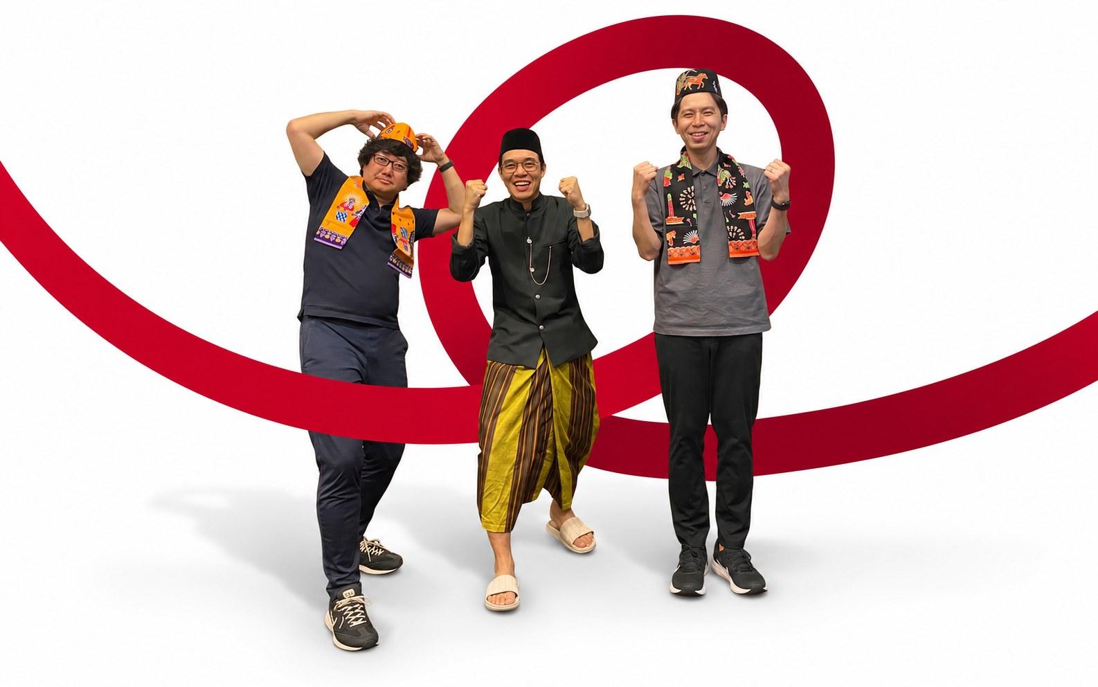 Two Japanese colleagues wearing songkok caps and batik sashes pose with an Indonesian colleague in traditional dress, fists raised together, with a looping brand ribbon sweeping behind them.