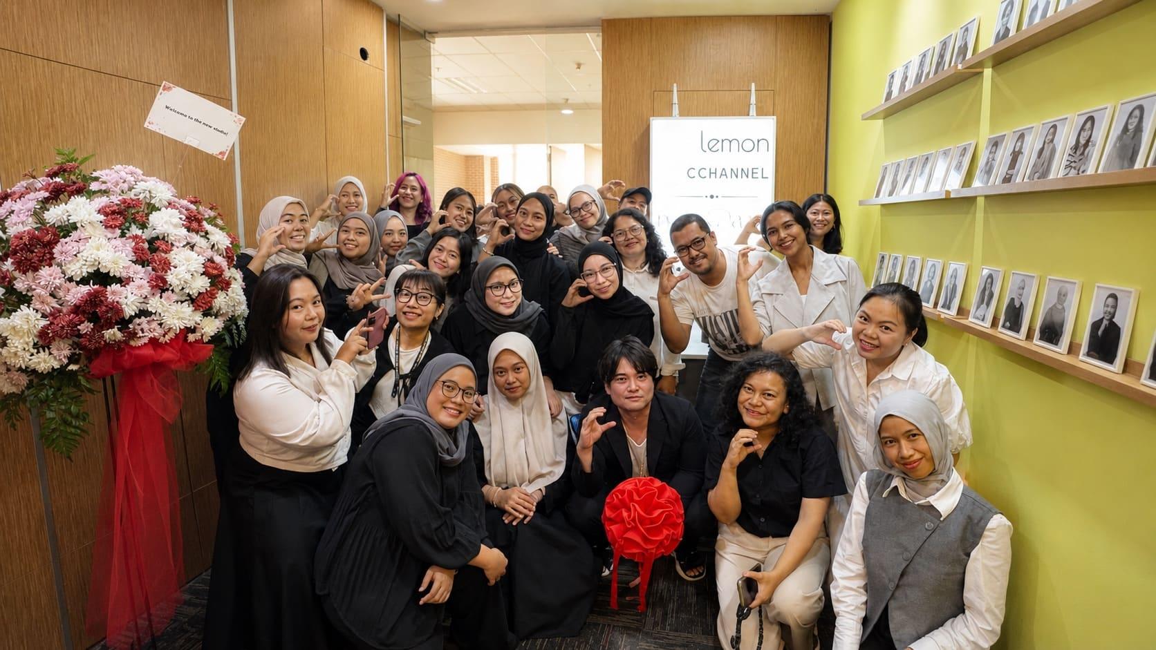 The C CHANNEL and lemon team packed together in the new Mega Kuningan office on opening day, a congratulatory flower stand by the door and the staff portrait wall behind them.