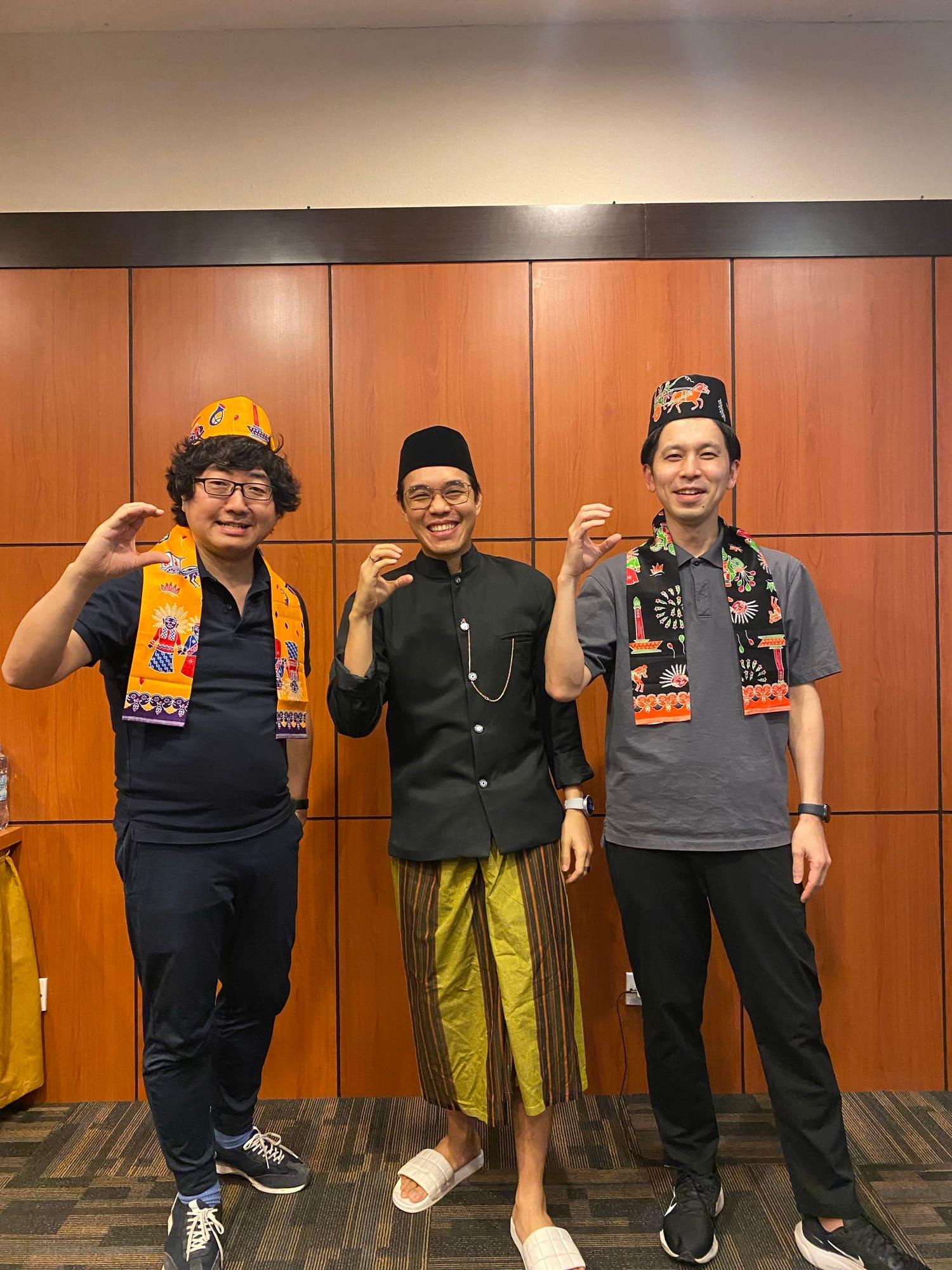 Three colleagues in Betawi caps and batik sashes strike a pose against the wood-panelled wall of a meeting room on a traditional dress day.