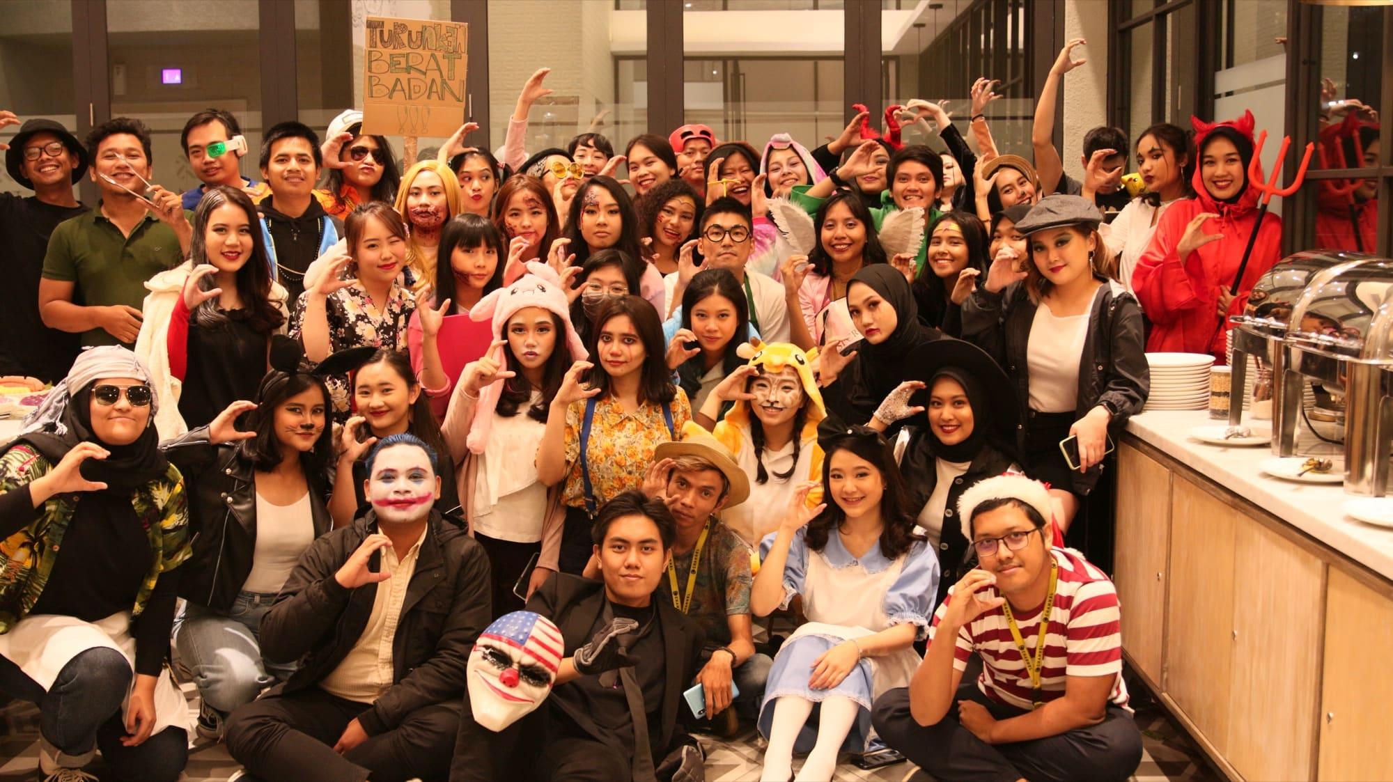 A large costume party: dozens of colleagues in face paint, wigs, animal hoods and hats crowd together for a group photo beside a buffet counter.