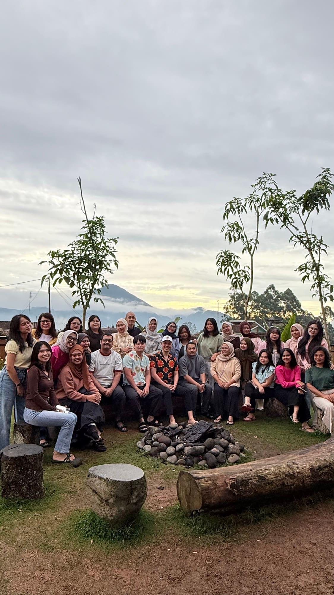The whole team gathered around a stone firepit at a hillside retreat at dawn, mountains and low cloud behind them.
