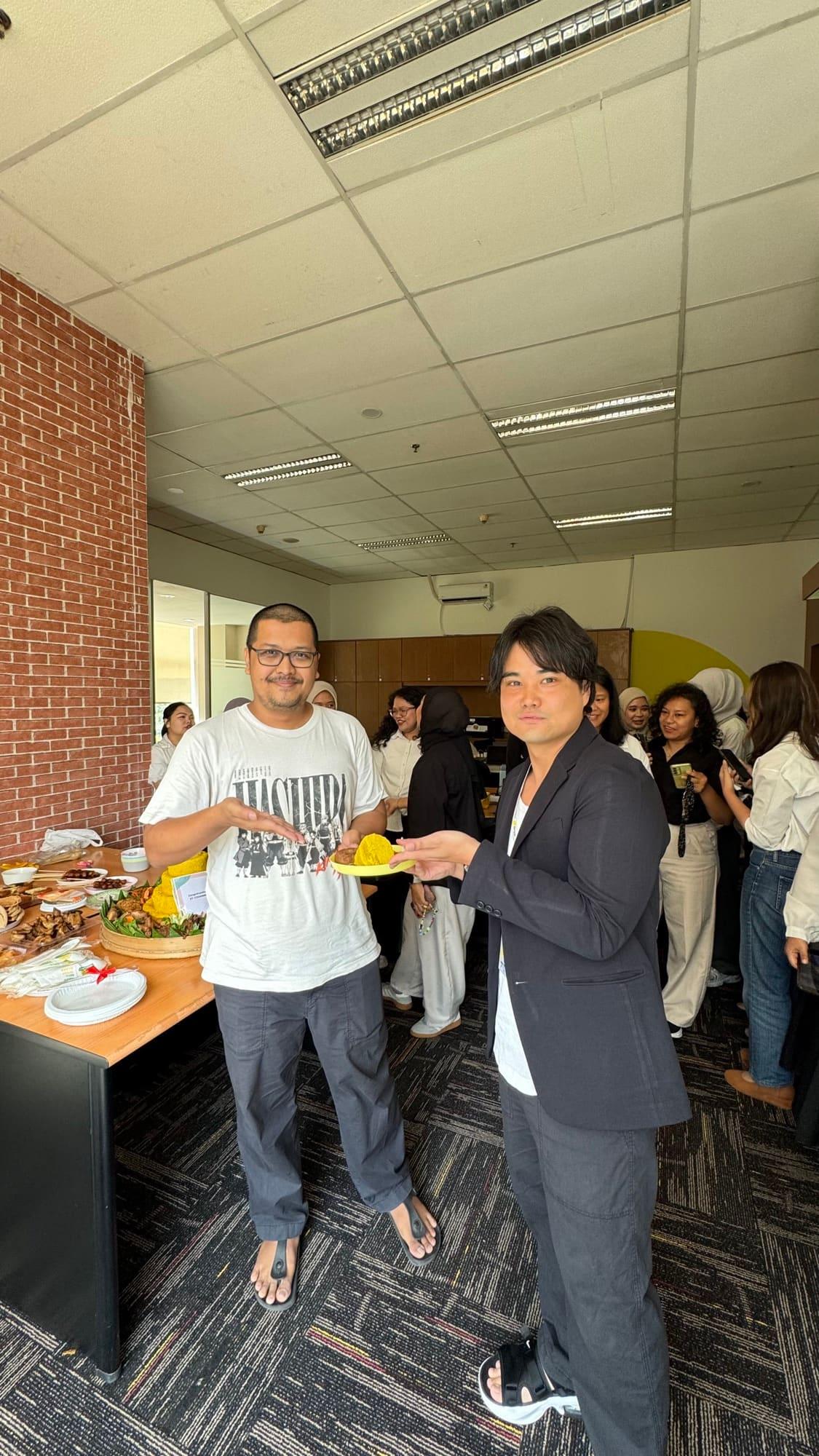 Two colleagues present a tumpeng, the yellow rice cone served at Indonesian celebrations, to the office over a table of shared dishes.