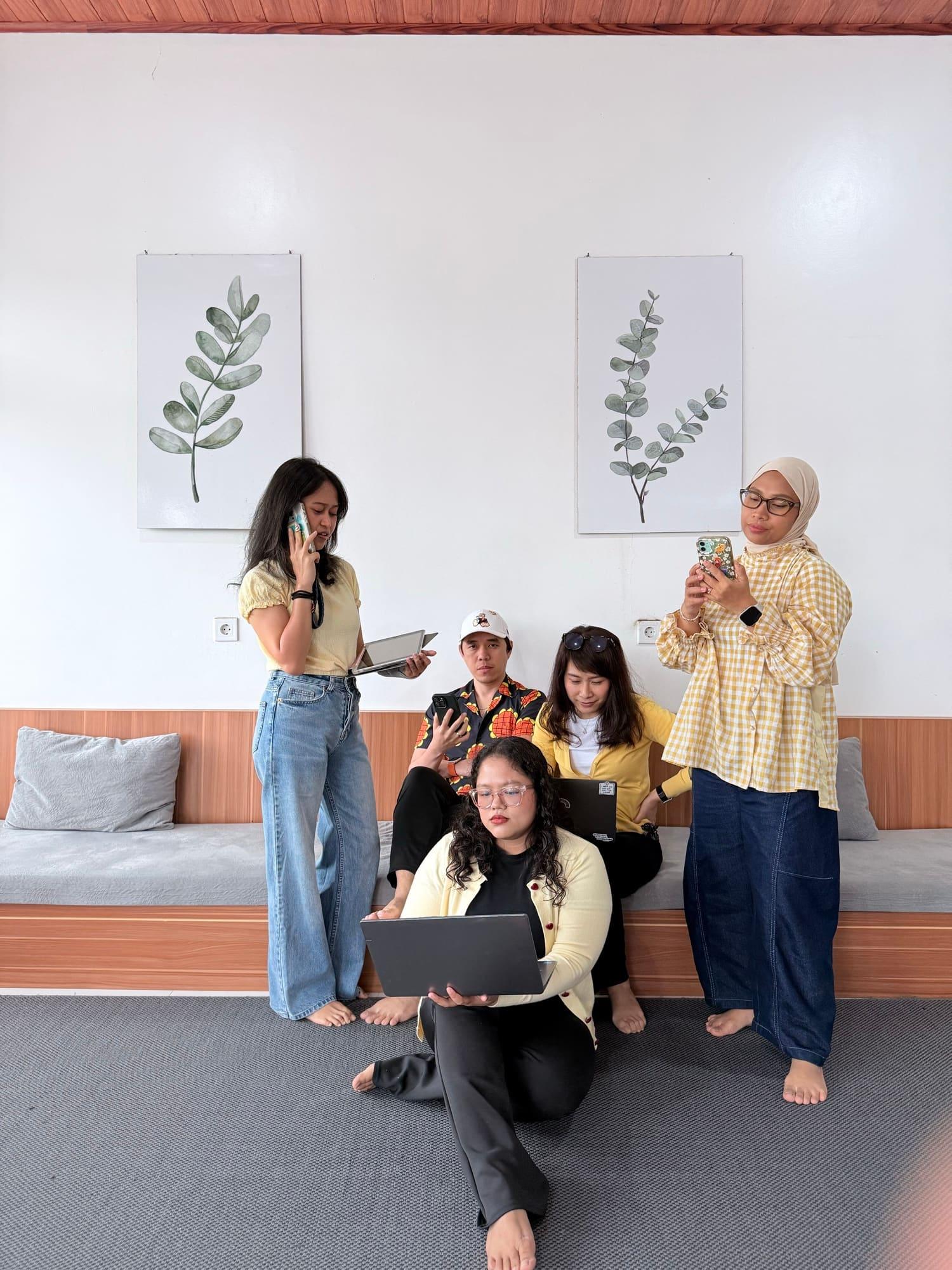 Five colleagues working from a villa living room during an offsite, laptops and phones in hand, sitting on the floor and a low bench.
