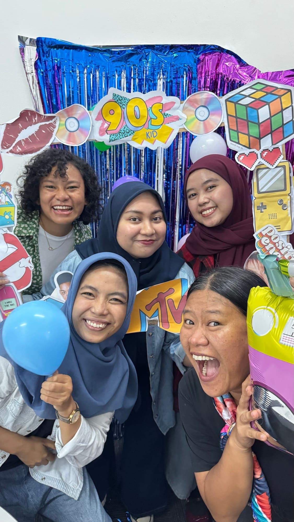 Five colleagues squeeze into a nineties-themed photo booth hung with foil fringe, holding balloons and cut-out props.