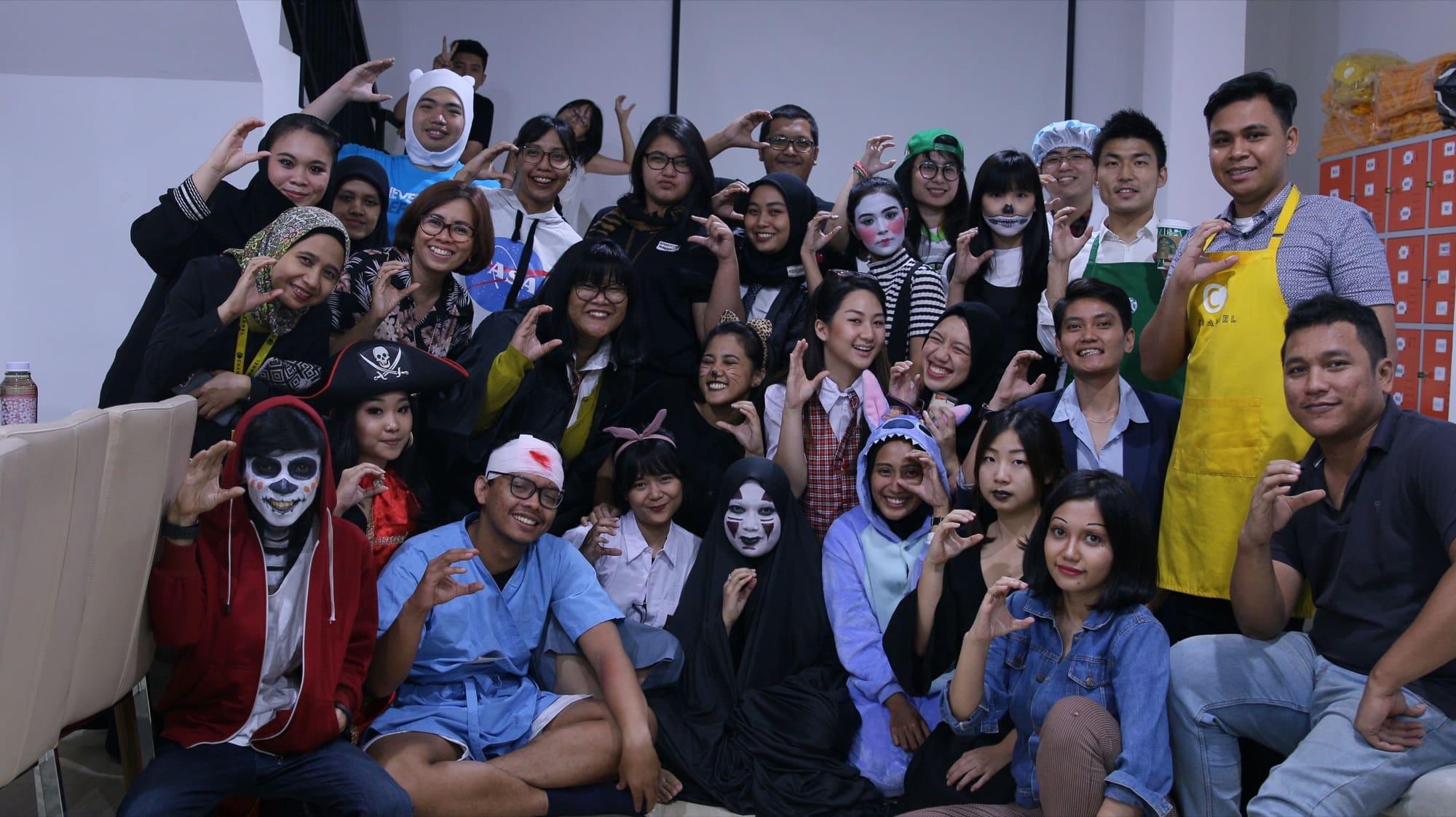 An earlier costume party at the old office, the team in face paint and fancy dress around the lockers, one in a C CHANNEL apron.