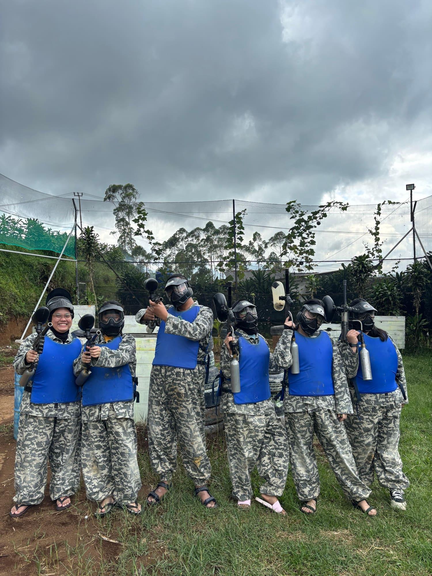 Seven colleagues in camouflage overalls and body armour line up with paintball markers on a field under a heavy sky.
