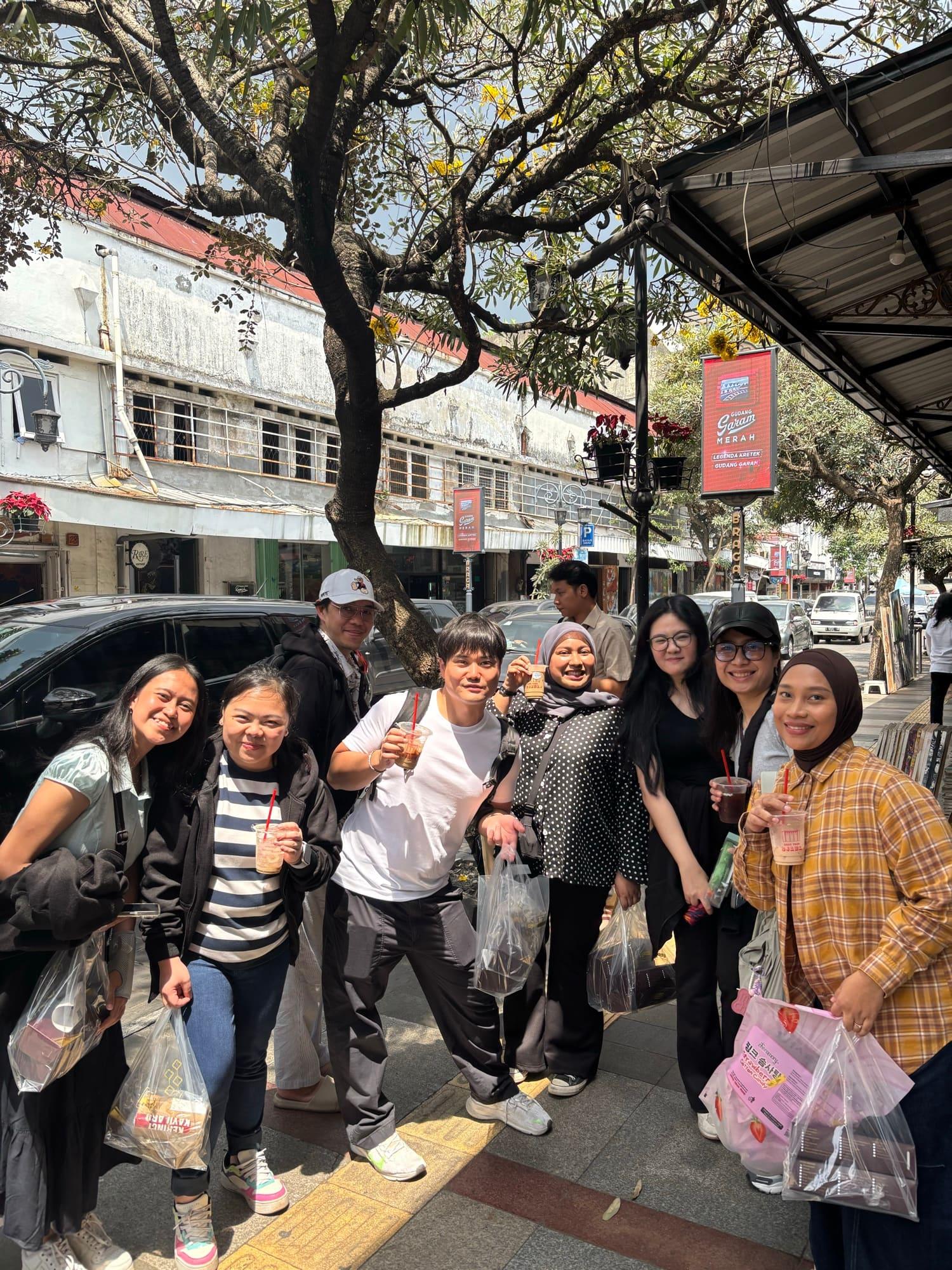 The team out on a colonial-era shopping street on a company trip, iced drinks and shopping bags in hand under a flowering tree.